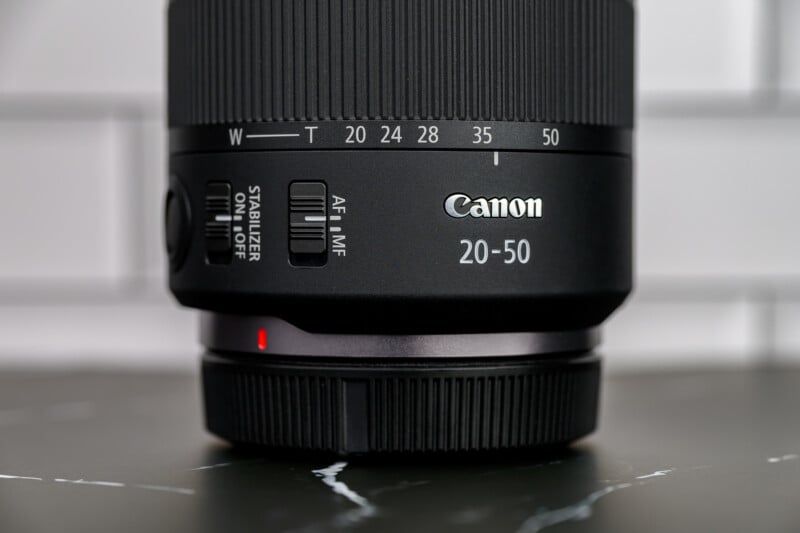 Close-up of a Canon camera lens showing the brand name, focal length numbers (20-50mm), and switches for image stabilizer and autofocus/manual focus, with a blurred white tiled background.