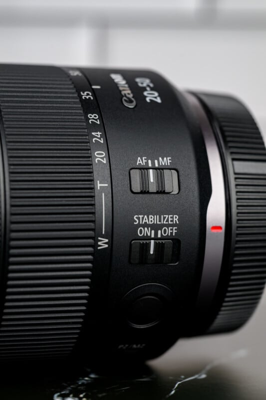 Close-up of a camera lens showing zoom markings, an AF/MF focus mode switch, and a stabilizer on/off switch. The lens has a ribbed grip and a visible red alignment dot.