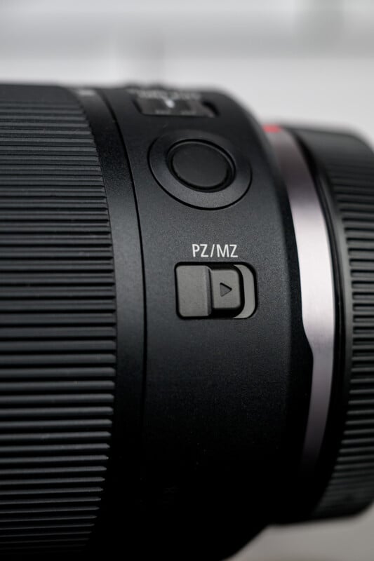 Close-up of a camera lens showing a textured focus ring, a large circular button, and a switch labeled "PZ/MZ" with a directional arrow. The lens is black with metallic accents.
