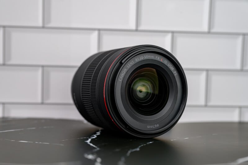 A Canon RF 20-50mm f/4 L lens resting on its side on a dark marble surface, with a white tiled wall in the background. The lens features a red ring near the front element.