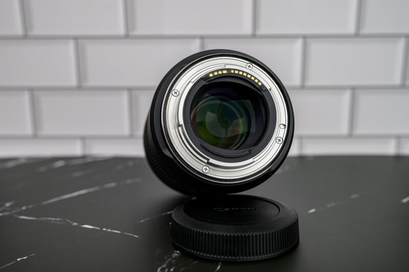 A camera lens lies face down on a black surface with its rear element and electronic contacts visible; the lens cap rests nearby. The background is a white subway tile wall.