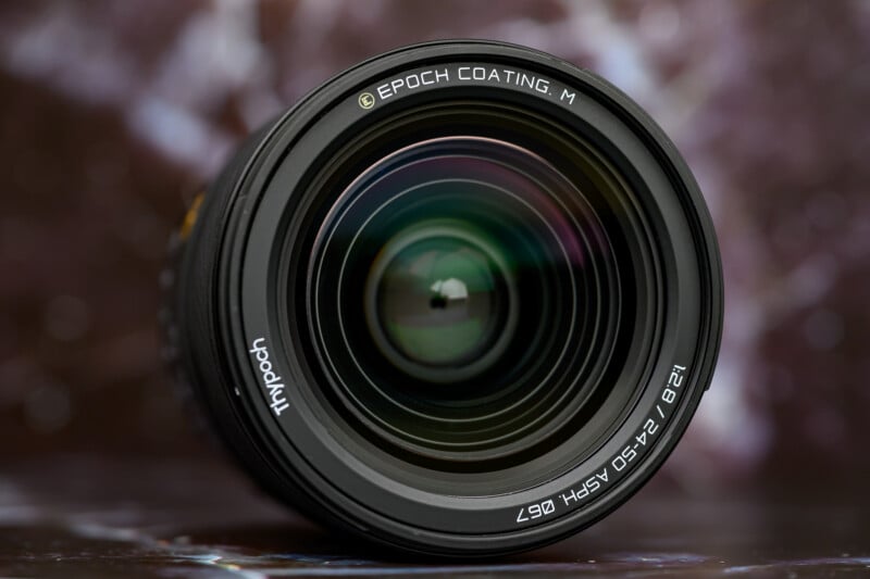 Close-up of a camera lens facing forward, showing detailed glass reflections and text around the rim reading "EPOCH COATING M" and "1:2.8 17-50 ASPH Ø67." The background is blurred with purple and white tones.