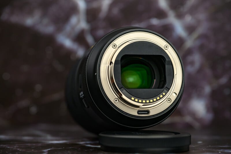 A close-up image of a camera lens resting on its side with the lens mount facing forward, showing the metal contacts and glass elements, with a lens cap nearby on a marble surface.