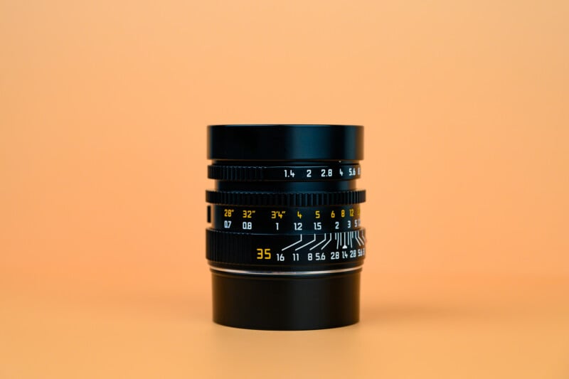 A black camera lens with white and yellow markings is standing upright on an orange surface, with a matching orange background.