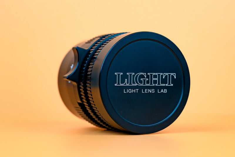 A black camera lens with the words "LIGHT LIGHT LENS LAB" printed on the front cap, placed on its side against a solid light orange background.