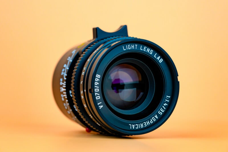 A close-up of a camera lens labeled "Light Lens Lab 1:1.2/35 ASPH EIRCIAL VI 070198" against a smooth peach-colored background. The lens is positioned upright, facing the viewer.