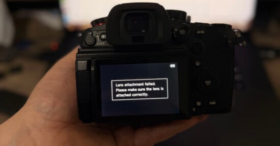 A hand holds a camera with its screen displaying an error message: "Lens attachment failed. Please make sure the lens is attached correctly.