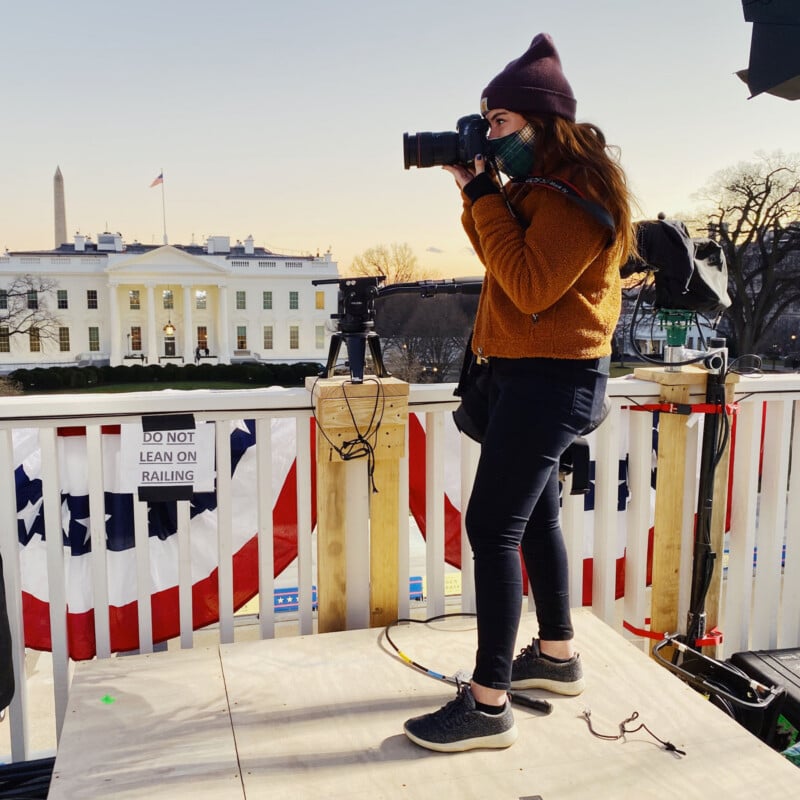 A woman wearing a beanie and mask stands on a platform taking photos with a camera. The White House and the Washington Monument are visible in the background at sunset. A sign reads, “DO NOT LEAN ON RAILING.”.