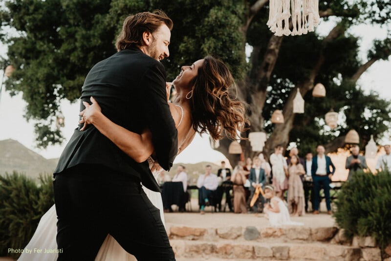 A bride and groom joyfully dance outdoors under trees, surrounded by hanging lanterns and guests watching in the background. The couple smiles, embracing each other in the center of a rustic wedding celebration.