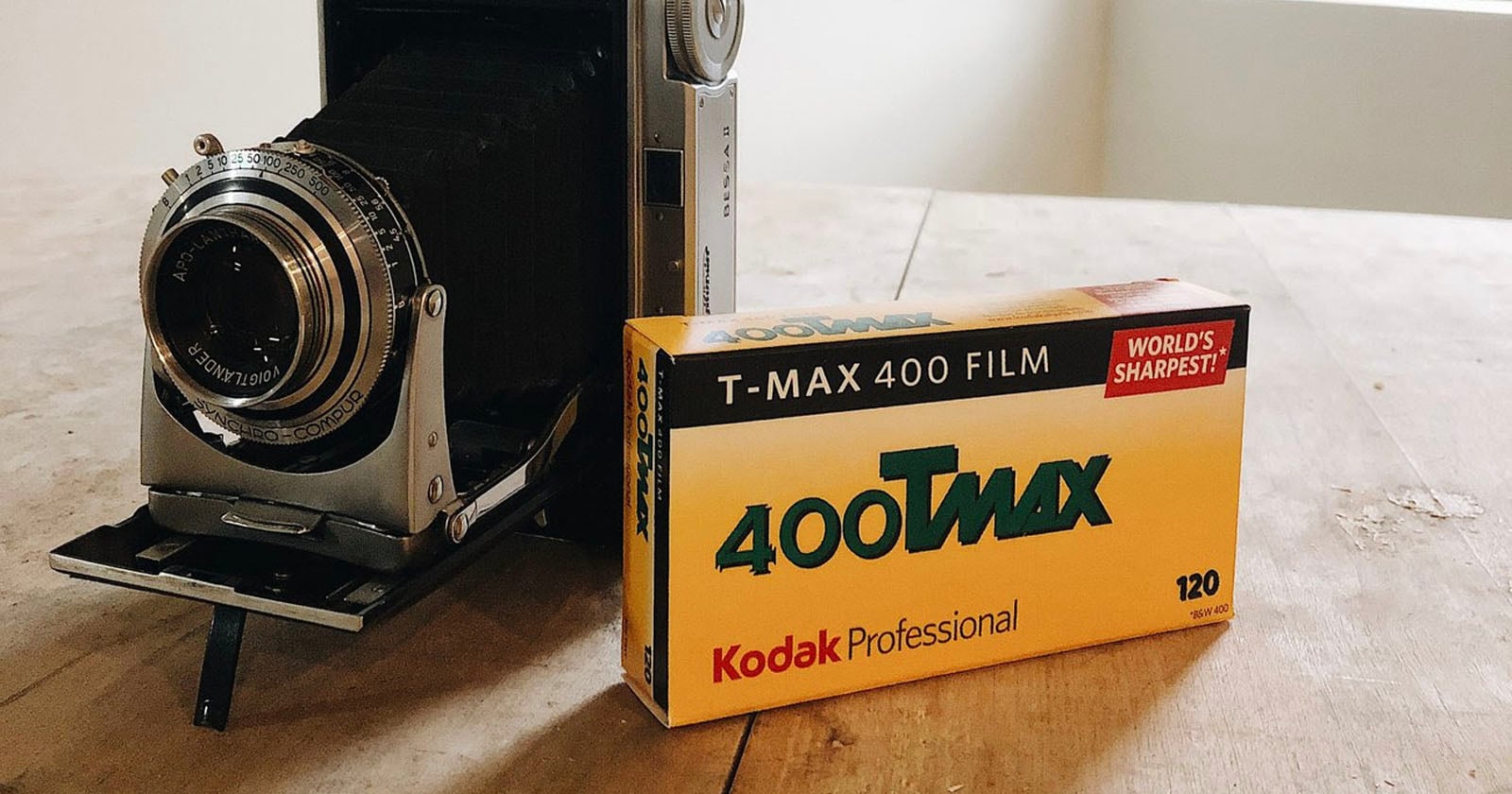 A box of Kodak Professional 400 T-Max 120 film sits on a wooden table next to a vintage camera. The film box is yellow with black and green text.