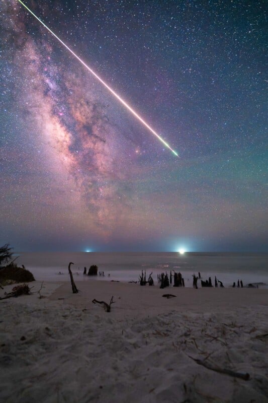 A bright meteor streaks across a star-filled night sky with the Milky Way visible, above a sandy beach with scattered driftwood and calm ocean waves illuminated by distant lights.