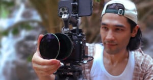 A person wearing a backward cap adjusts a filter on a professional camera mounted on a tripod outdoors, with a smartphone attached to the camera rig.