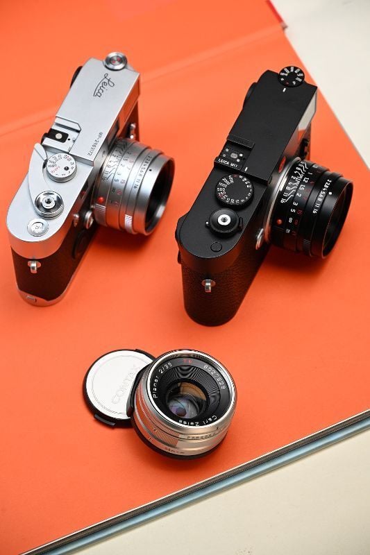 Two vintage cameras with attached lenses and a detached lens with its cap are displayed on a bright orange surface. The cameras are positioned at an angle, showcasing their top dials and controls.