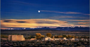 A small village with adobe buildings sits under a colorful sunset sky, with the moon visible above distant snow-capped mountains and autumn trees in the background.