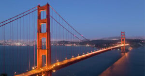 The Golden Gate Bridge illuminated at dusk, stretching across the water with the city skyline visible in the background under a clear, blue evening sky.