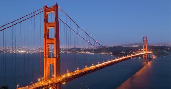 The Golden Gate Bridge illuminated at dusk, stretching across the water with the city skyline visible in the background under a clear, blue evening sky.