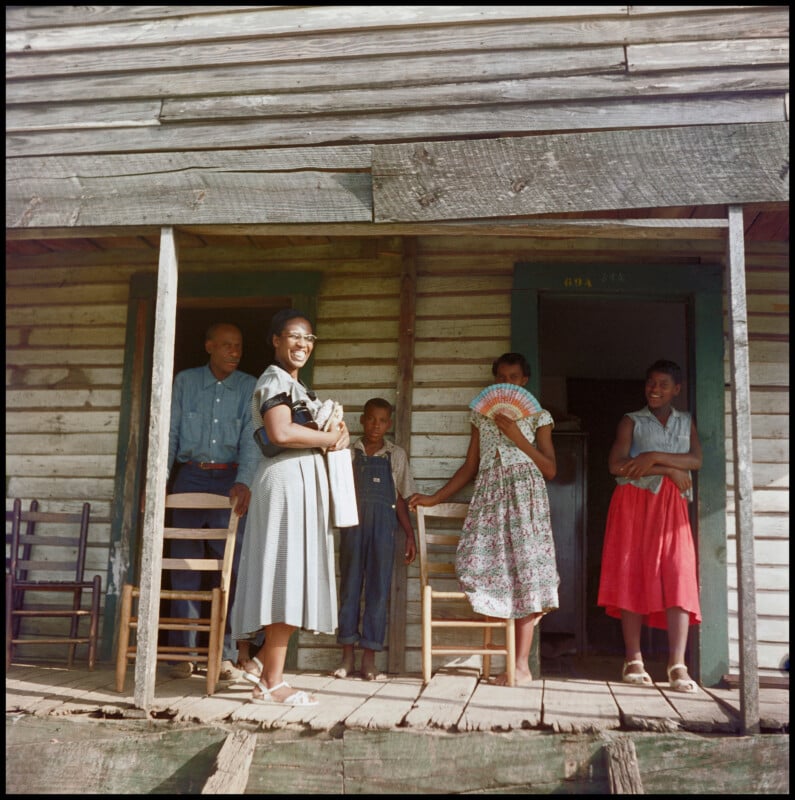 Five people stand and sit on a rustic wooden porch. A woman smiles at the camera, while others, including children, stand or sit nearby. The house has weathered wood siding and simple furnishings.
