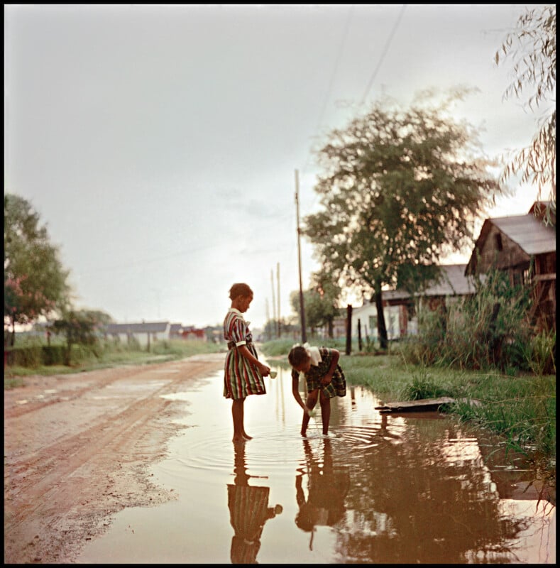 Two young girls stand barefoot in a large puddle on a dirt road; one bends down to touch the water while the other looks on. Trees, grass, and houses line the road under a cloudy sky.