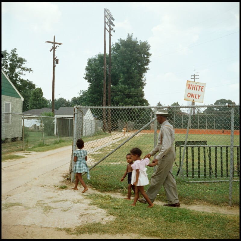 A Black man holds hands with three young Black girls as they walk past a chain-link fence with a sign that reads "WHITE ONLY." The scene is outdoors near a grassy field and utility poles.