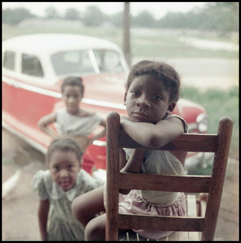 Three young Black girls stand and sit outside near a wooden chair; one leans her arms on the chair's back, looking quietly at the camera. Behind them is a vintage red and white car and a blurred green landscape.