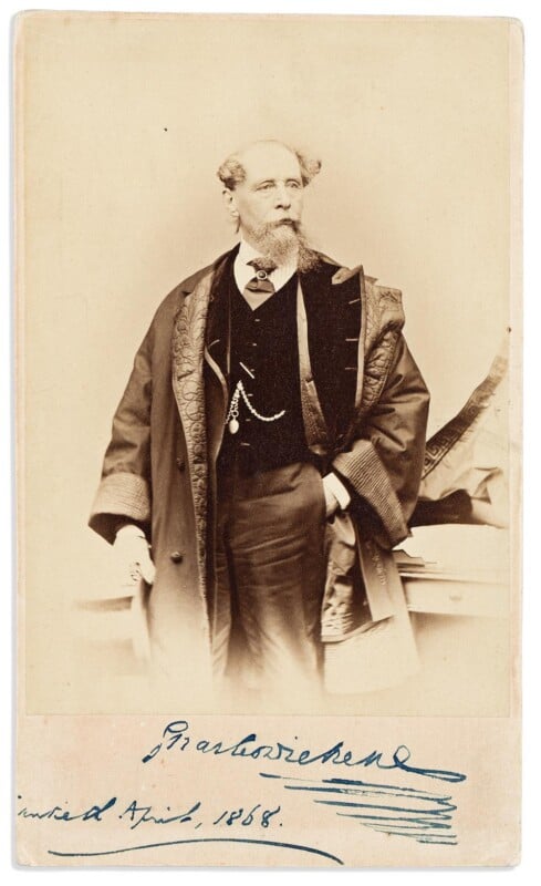 A Victorian-era man with a beard stands in formal attire, wearing a long coat with ornate trim. One hand is tucked into his coat, and there is a handwritten signature at the bottom of the sepia-toned photograph.