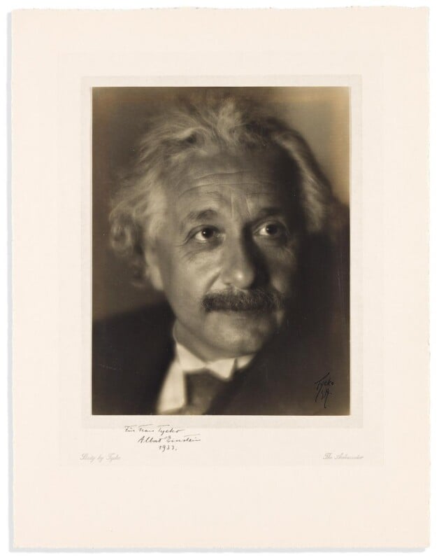 A sepia-toned portrait of an older man with wavy white hair and a mustache, wearing a suit and looking slightly to the side against a soft, blurred background. Handwritten notes and a signature appear below the image.