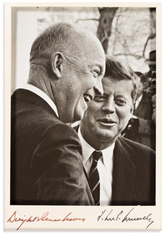 Two men in suits stand close together, smiling and talking outdoors. The photo is black and white, with handwritten signatures beneath the image. A blurred crowd and trees are visible in the background.