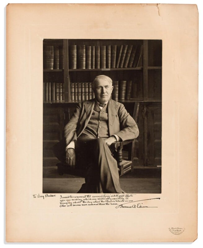 A black-and-white photo of Thomas Edison seated in a chair, wearing a suit, with bookshelves behind him. The photo is mounted on a mat with a handwritten note and signature at the bottom.