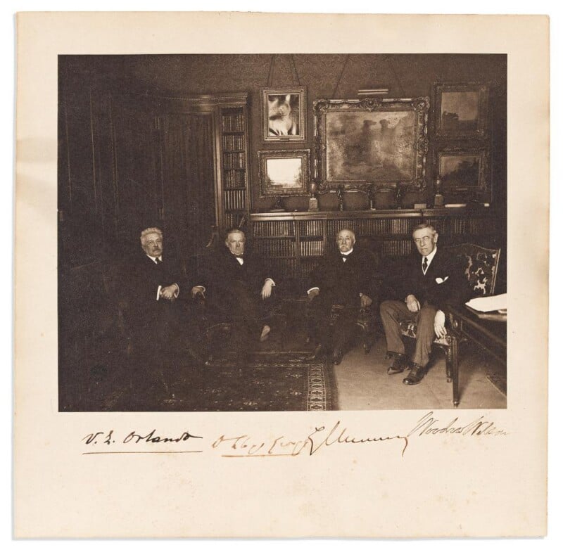 Four men in suits sit in an elegant, book-lined room with framed paintings on the wall. The men are facing the camera, seated on chairs and a sofa, with a rug on the floor and a large desk at the right.