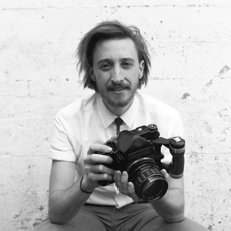 A person with medium-length hair and a mustache sits against a textured wall, wearing a short-sleeved button-up shirt and holding a medium format camera, smiling gently at the camera.