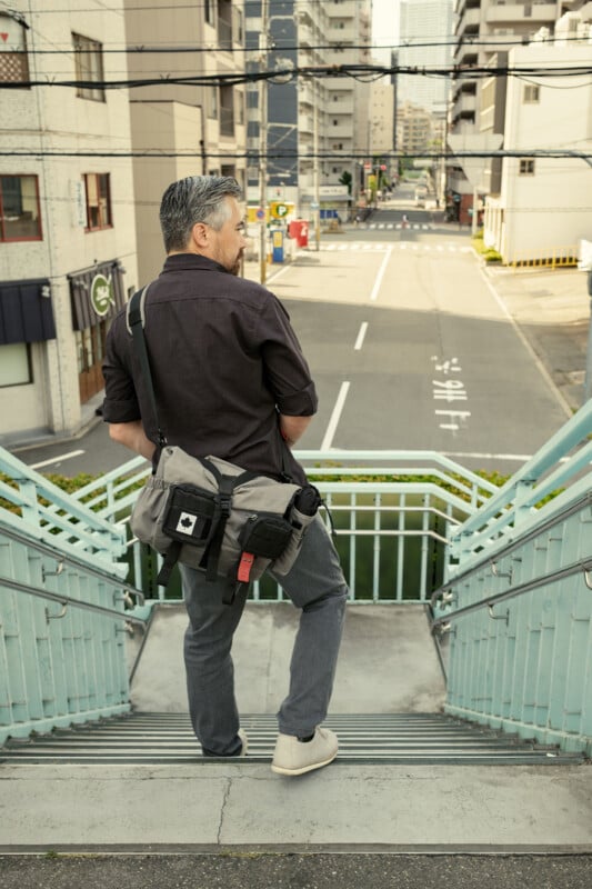 A man with gray hair stands on outdoor stairs, facing away, looking at a quiet urban street lined with tall buildings. He carries a gray messenger bag and wears casual clothes.