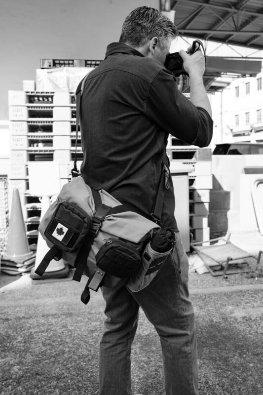 A person with a shoulder bag featuring a Canadian flag patch is taking photographs in an industrial area with stacked bricks and construction materials. The image is in black and white.