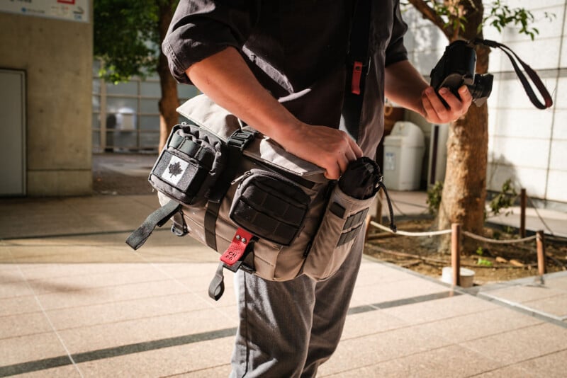 A person holding a camera in one hand and carrying a gray tactical shoulder bag with various pouches, standing outdoors on a sunlit walkway near a building and trees.