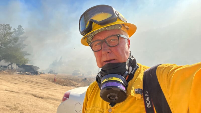 A firefighter in yellow protective gear and helmet takes a selfie at a smoky outdoor scene, with fire equipment and vehicles visible in the hazy background.