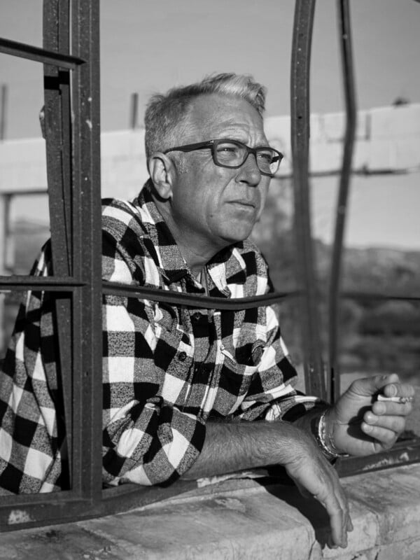 A man with light hair and glasses, wearing a checkered shirt, leans on a windowsill and looks outside thoughtfully while holding a cigarette in his hand. The window frame is metal and partially damaged. The image is black and white.