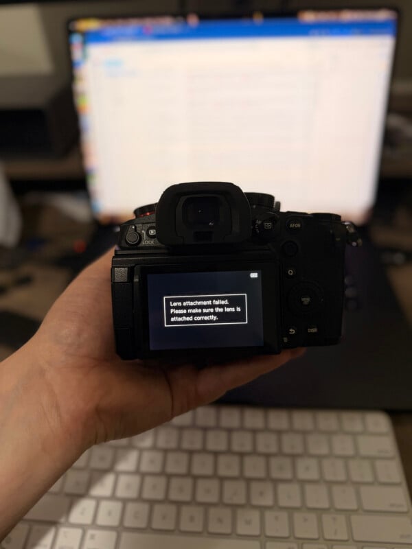 A hand holds a digital camera displaying an error message on its screen that reads, "Lens attachment failed. Please make sure the lens is attached correctly." A blurred computer monitor and keyboard are in the background.
