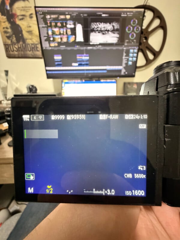 A close-up of a digital camera's display screen showing manual settings, with video editing software and a movie reel visible on two monitors in the blurred background. A "Rushmore" film poster is on the wall.