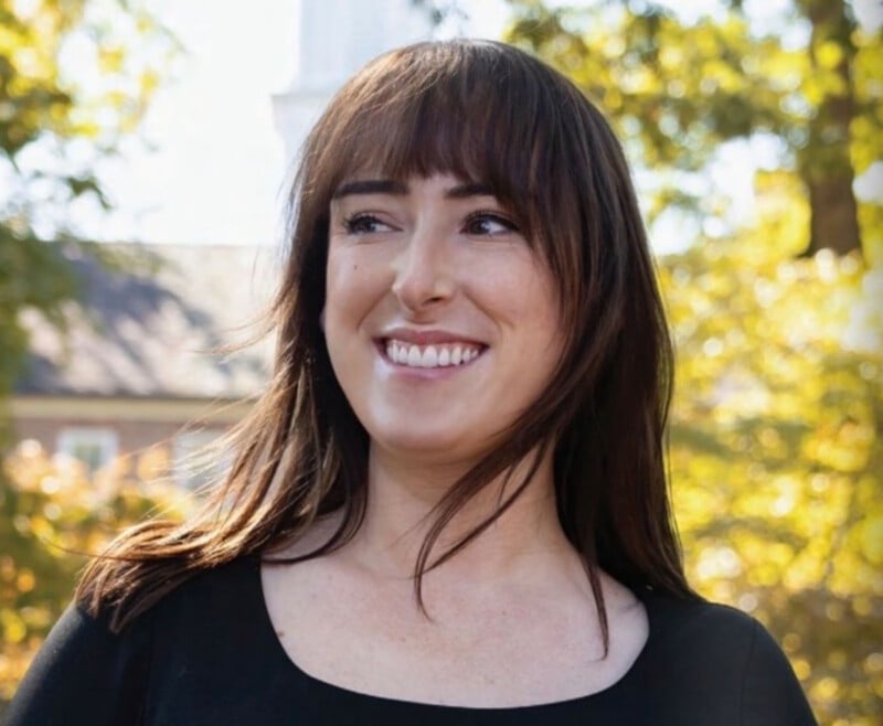 A woman with long brown hair and bangs smiles while standing outdoors in sunlight, with trees and a blurred building in the background. She is wearing a black top.