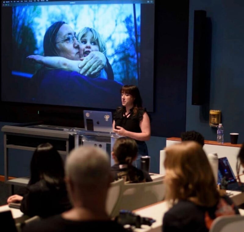 A woman gives a presentation to an audience in a lecture hall. Behind her, a large screen displays an image of a woman holding a child outdoors. The audience listens attentively.