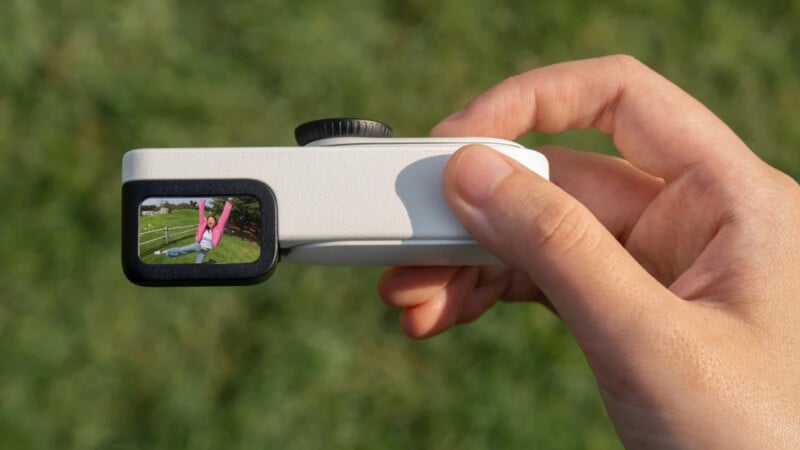 A hand holds a small, rectangular camera with a display screen showing a person in a pink shirt doing a handstand on grass, with a blurred green background.
