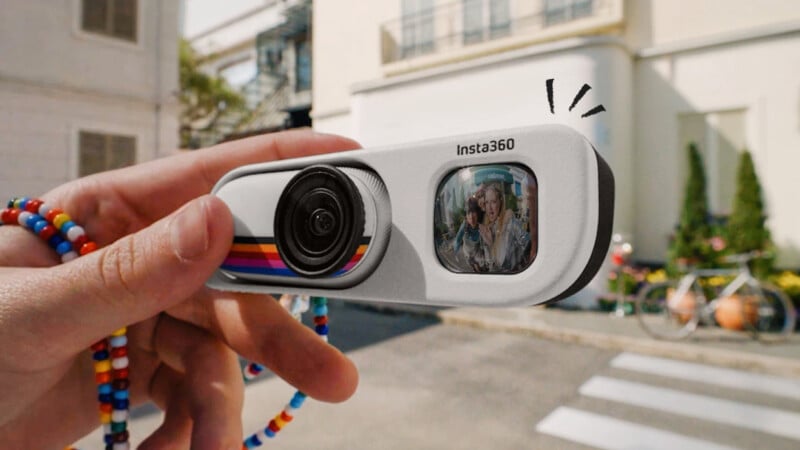 A hand holds an Insta360 camera with a beaded strap; the camera’s screen displays a person smiling outdoors, with a blurred building and street in the background.