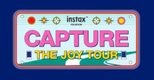 A colorful sign styled like a license plate reads "instax FUJIFILM CAPTURE THE JOY TOUR" with stars, circles, and a winding road graphic against a blue background.