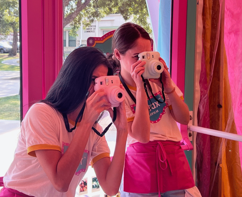 Two women in matching white and pink outfits hold instant cameras to their faces, taking photos inside a bright, colorful room with pink walls and large windows showing trees outside.