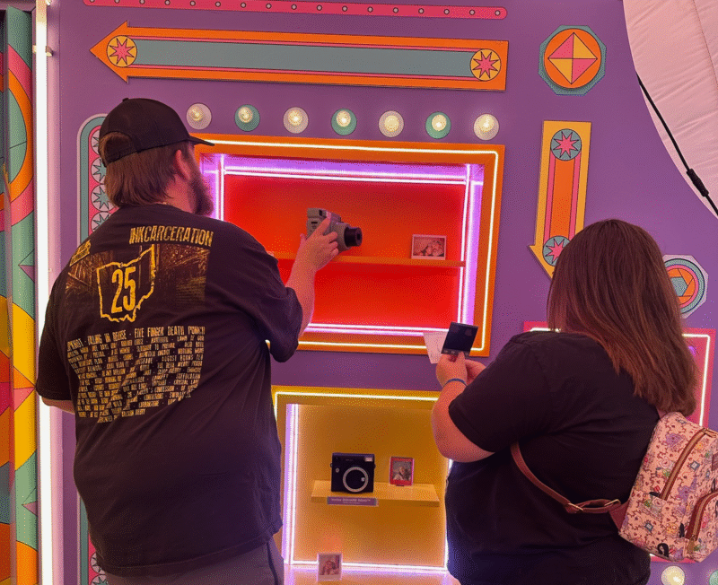 Two people stand in front of a brightly colored, neon-lit display wall, taking photos of instant cameras and small photos on shelves. One holds a camera while the other uses a phone. The scene is playful and vibrant.