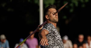 A man in a patterned shirt prepares to throw a javelin, with his arm extended and the javelin held near his face; blurred spectators are visible in the background.