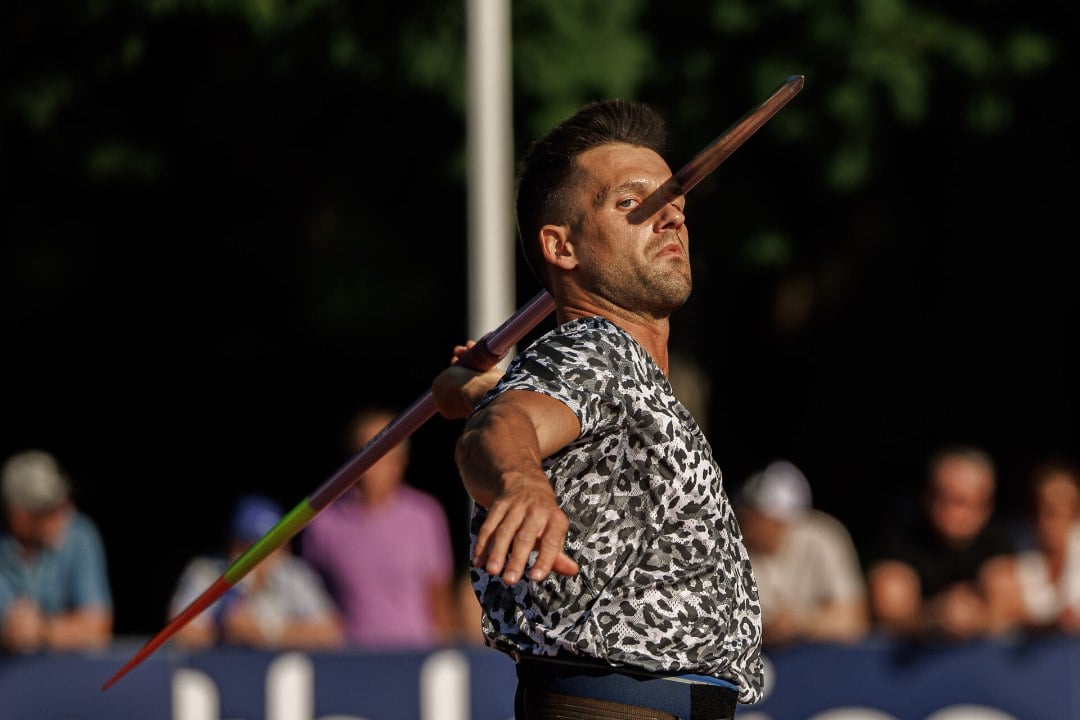 A male athlete in a patterned shirt prepares to throw a javelin, his arm extended and focused expression, with a blurred crowd and trees in the background.