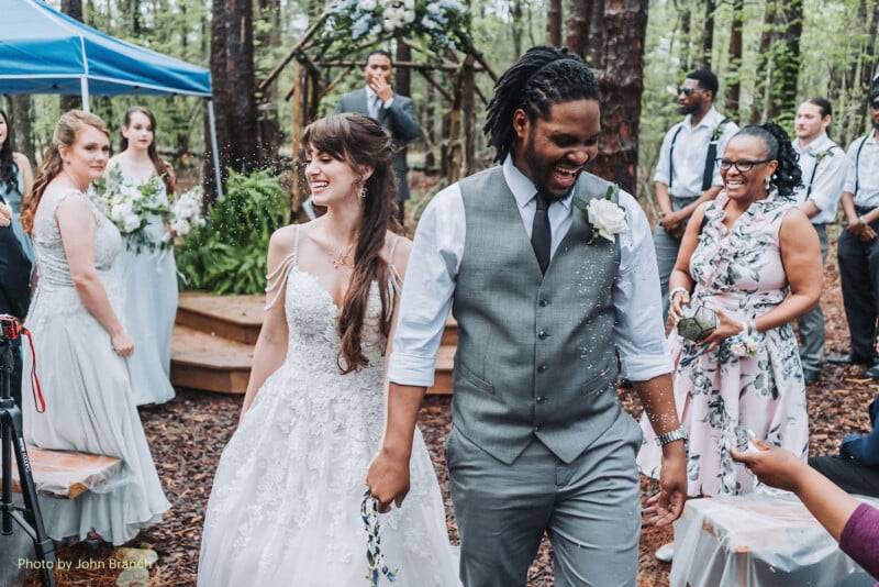 A bride and groom, smiling and holding hands, walk down the aisle at an outdoor forest wedding while guests throw confetti. Bridesmaids, groomsmen, and guests look on joyfully in the background.