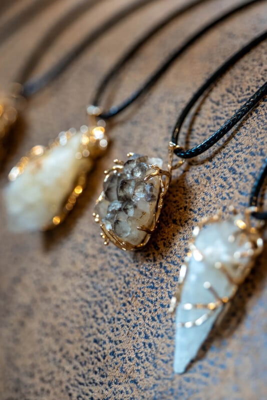 Three crystal pendants wrapped in gold wire are displayed on black cords against a speckled brown background. The crystals vary in shape and texture, including rough and pointed stones.