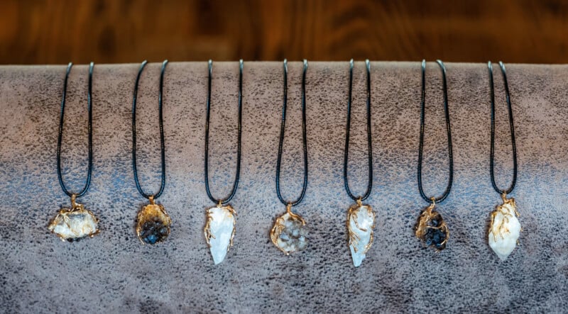Seven necklaces with black cords and wire-wrapped crystal pendants are displayed in a row on a textured gray surface, each featuring unique stone shapes and colors ranging from white to dark gray.