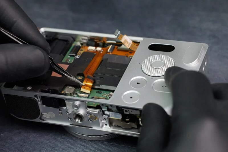 A person wearing black gloves uses tweezers to repair the internal components of a compact digital camera, exposing its circuit board and ribbon cables.